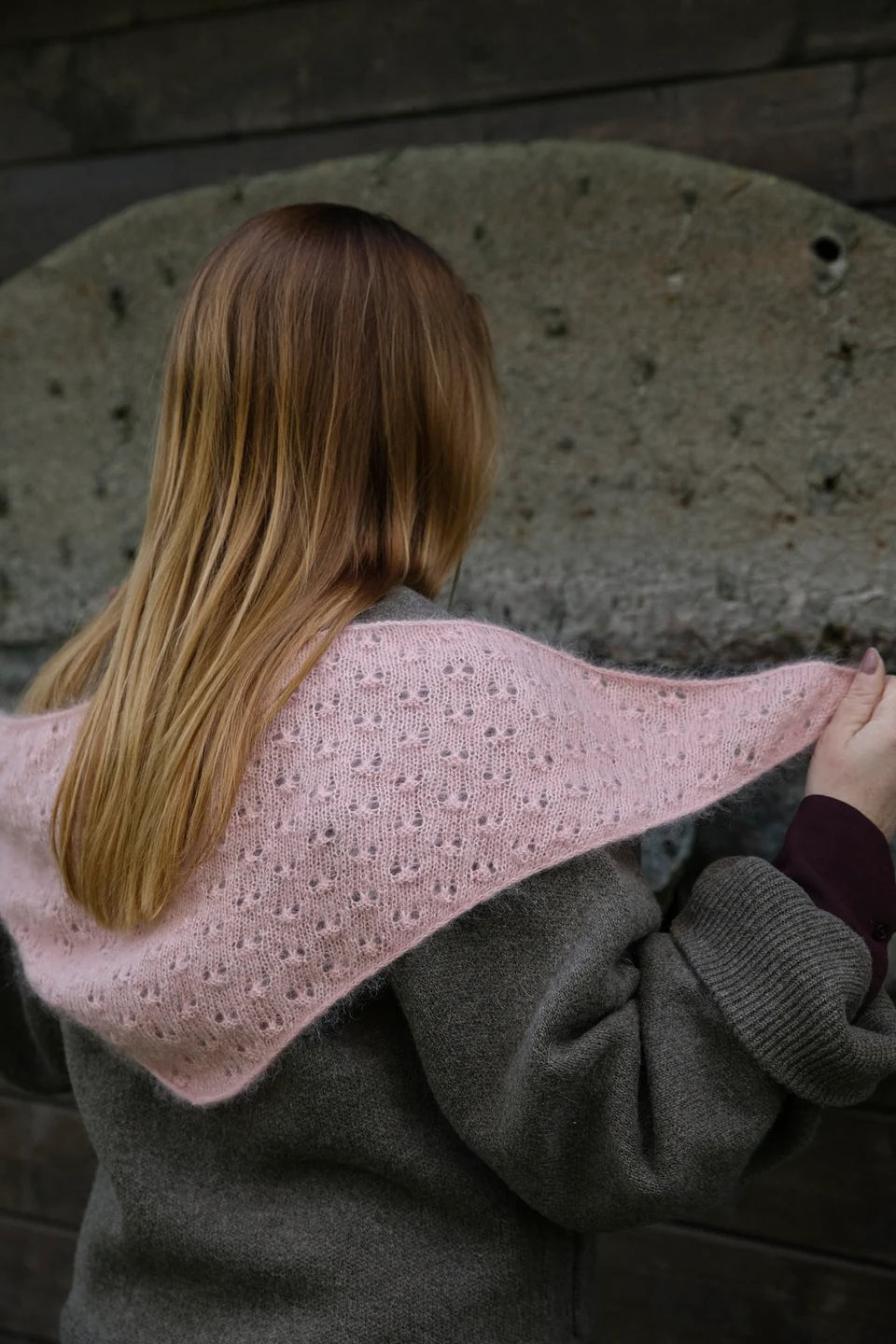 A person, viewed from behind, holding a pink lace knit shawl across their back, against a neutral background.