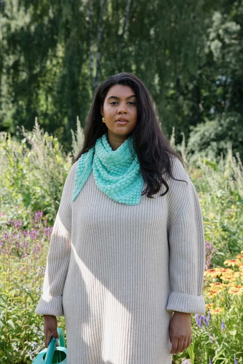 A person wearing an aqua knit shawl with bobbles on it around their neck standing in a natural outdoor setting.