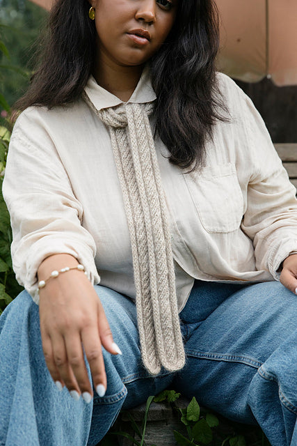 A person sitting down wearing a thin, beige knit scarf tied once around their neck.