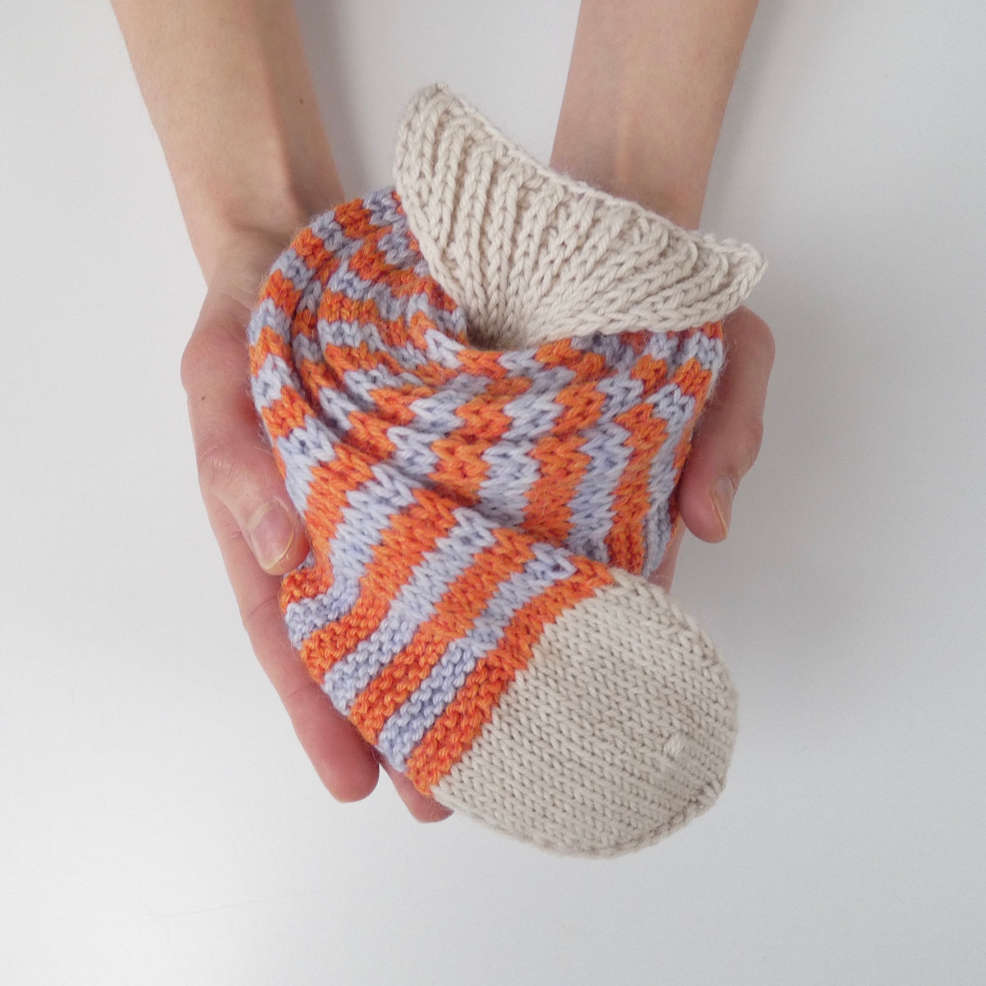 Person holding a rolled up knit skinny scarf in the shape of a fish with orange and light blue stripes on a white background.