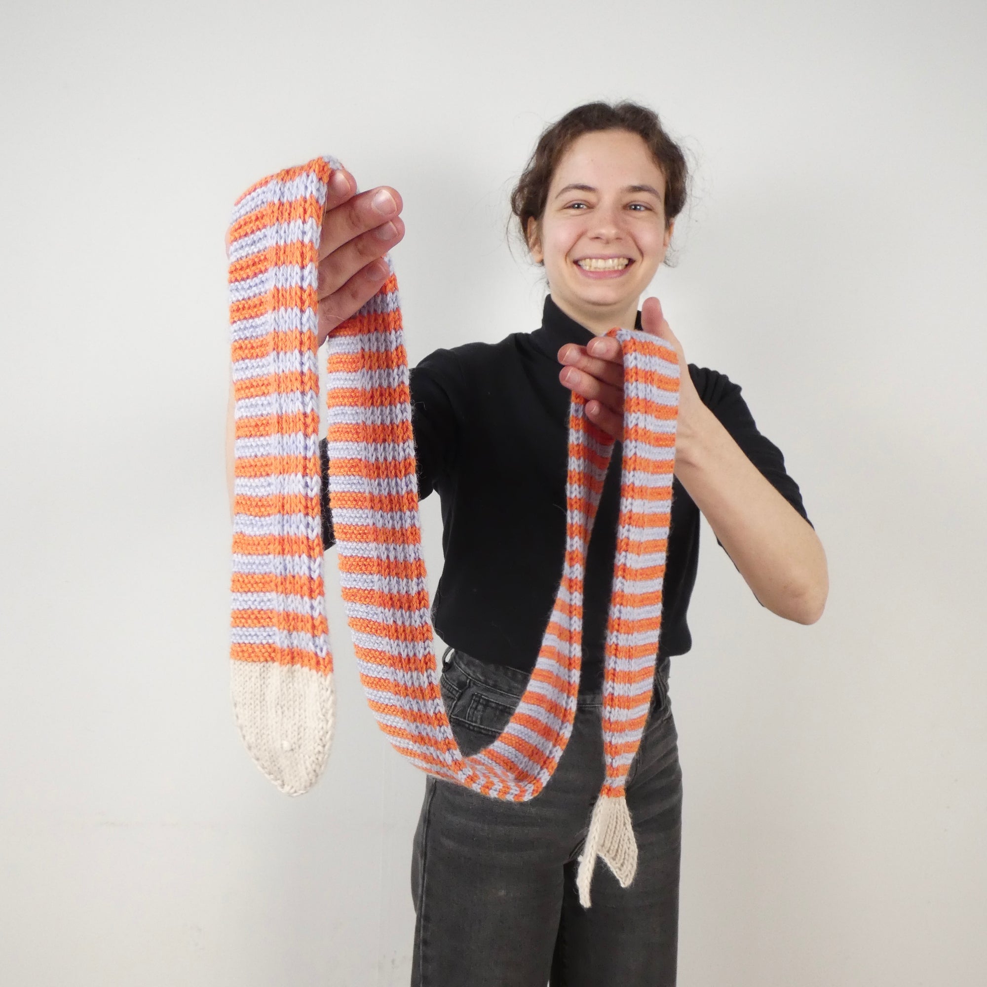 Person holding a striped knit skinny scarf in the shape of a fish with orange and light blue stripes against a plain background