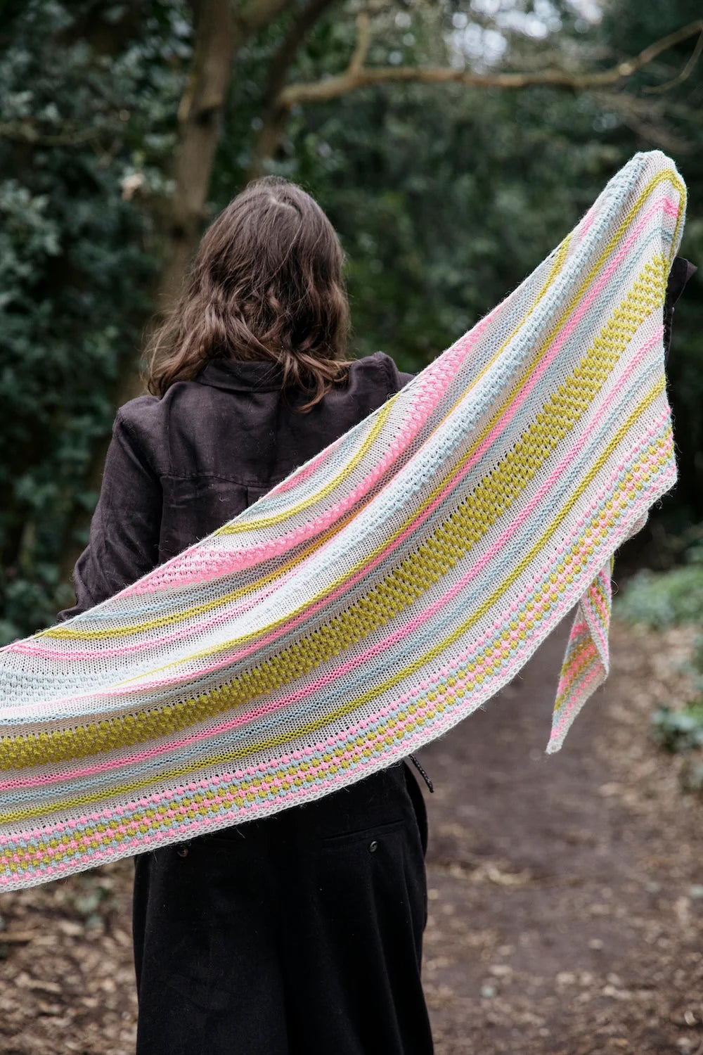 Person facing away and holding a colorful knit shawl across their back.