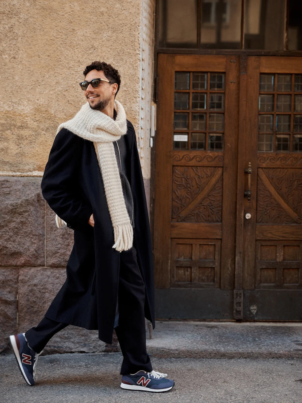 Man wearing a long coat, white knit scarf, and sunglasses walking on a street.