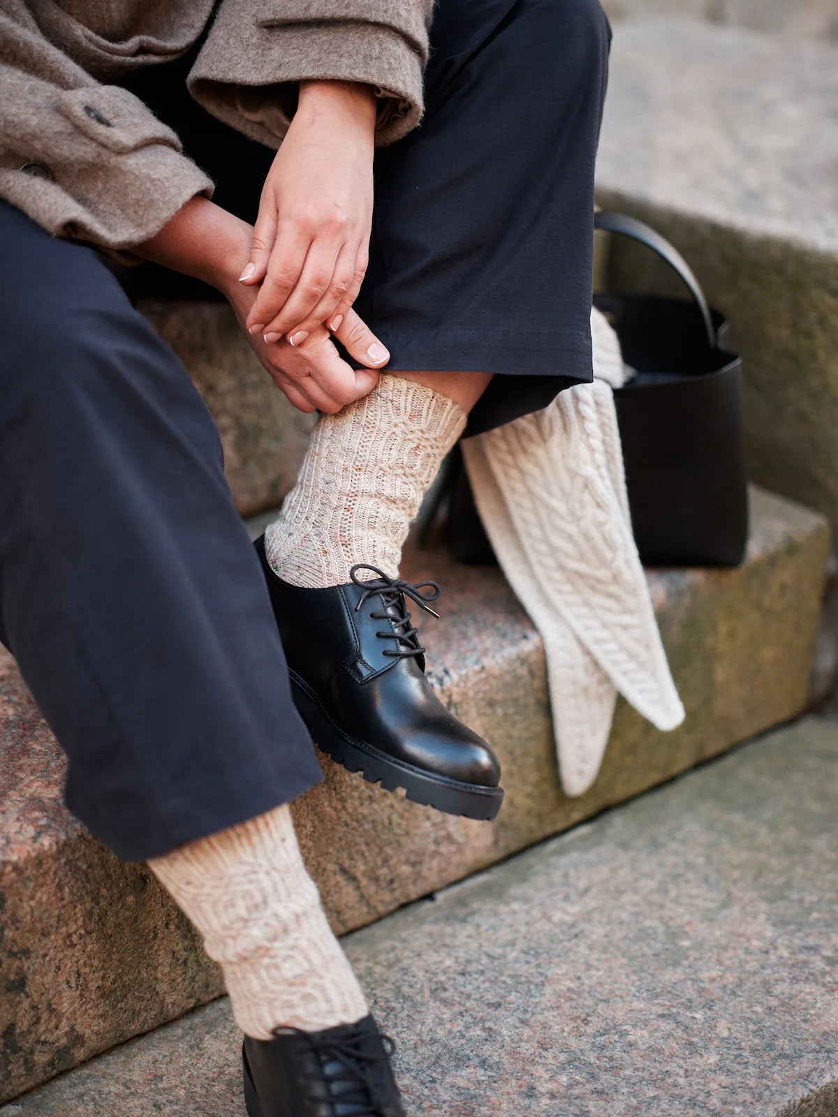 Person wearing black shoes and beige cable-knit socks sitting on a stone step.