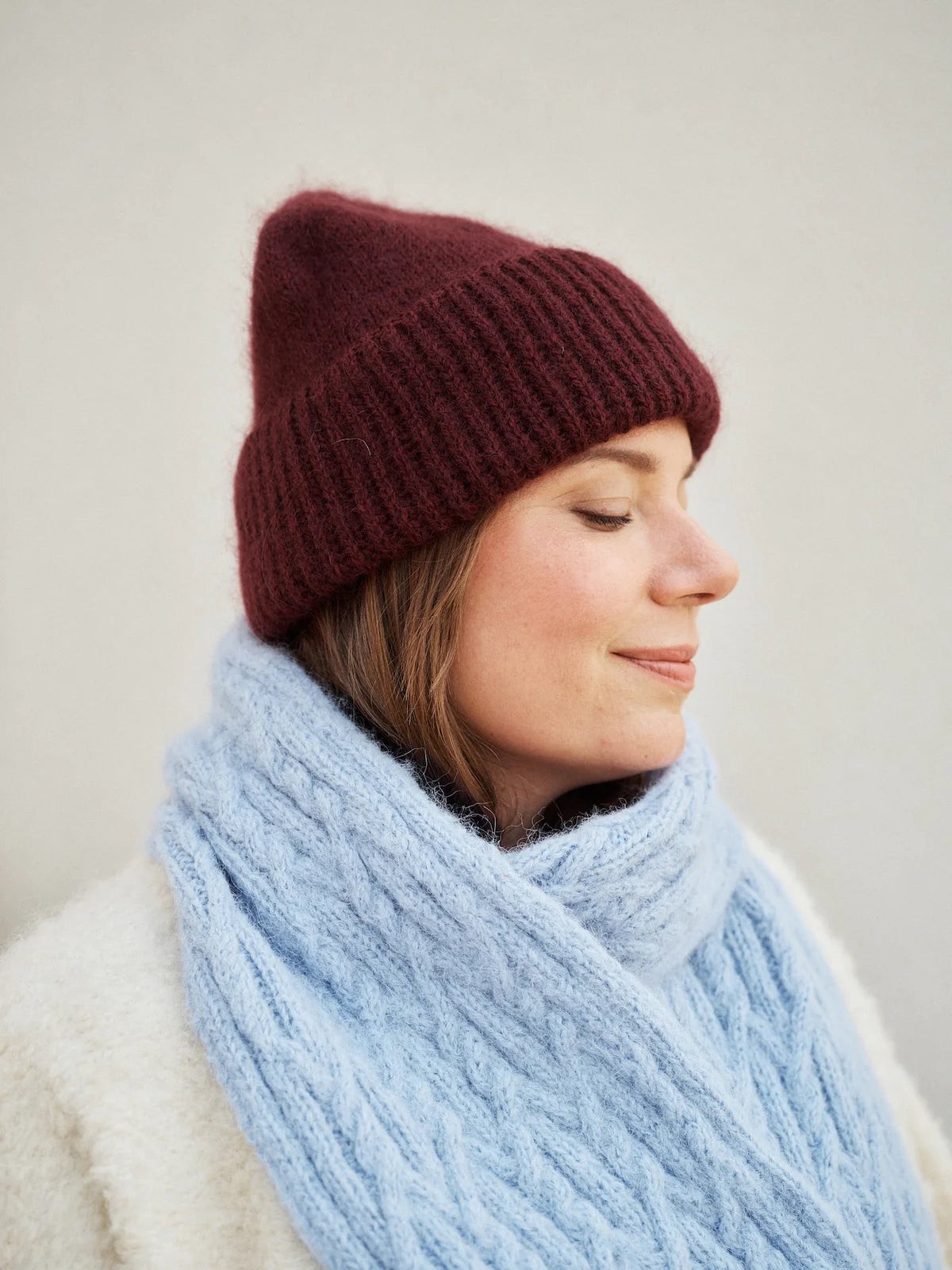 Woman wearing a burgundy knit beanie and light blue cable-knit scarf against a plain background