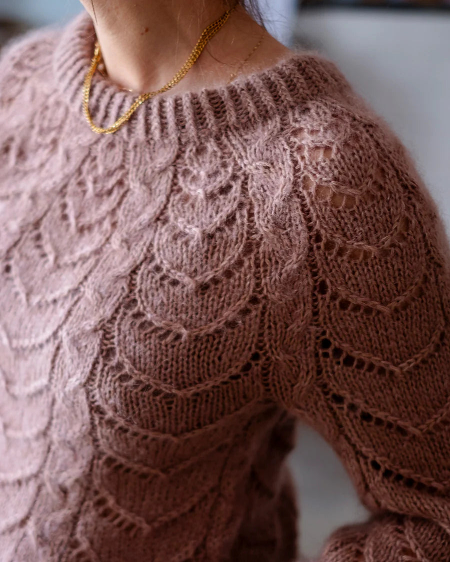 A close-up of a person wearing a dusty rose pink cabled and lace sweater, paired with a delicate gold chain necklace.