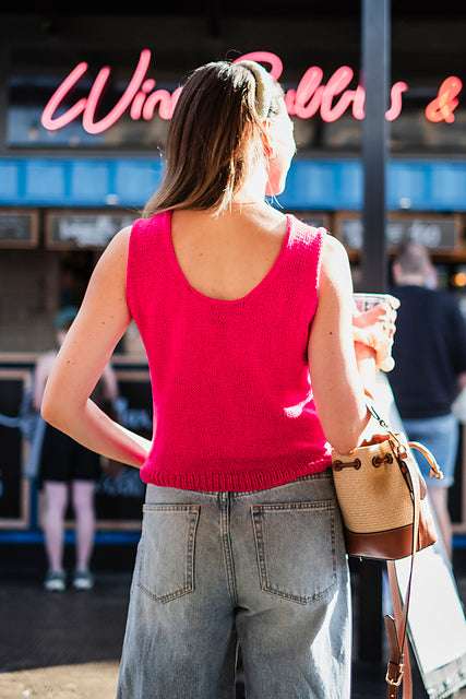 A woman viewed from behind wearing a bright pink sleeveless knit top and light-wash wide-leg jeans, standing outdoors in front of a storefront with a pink neon sign.