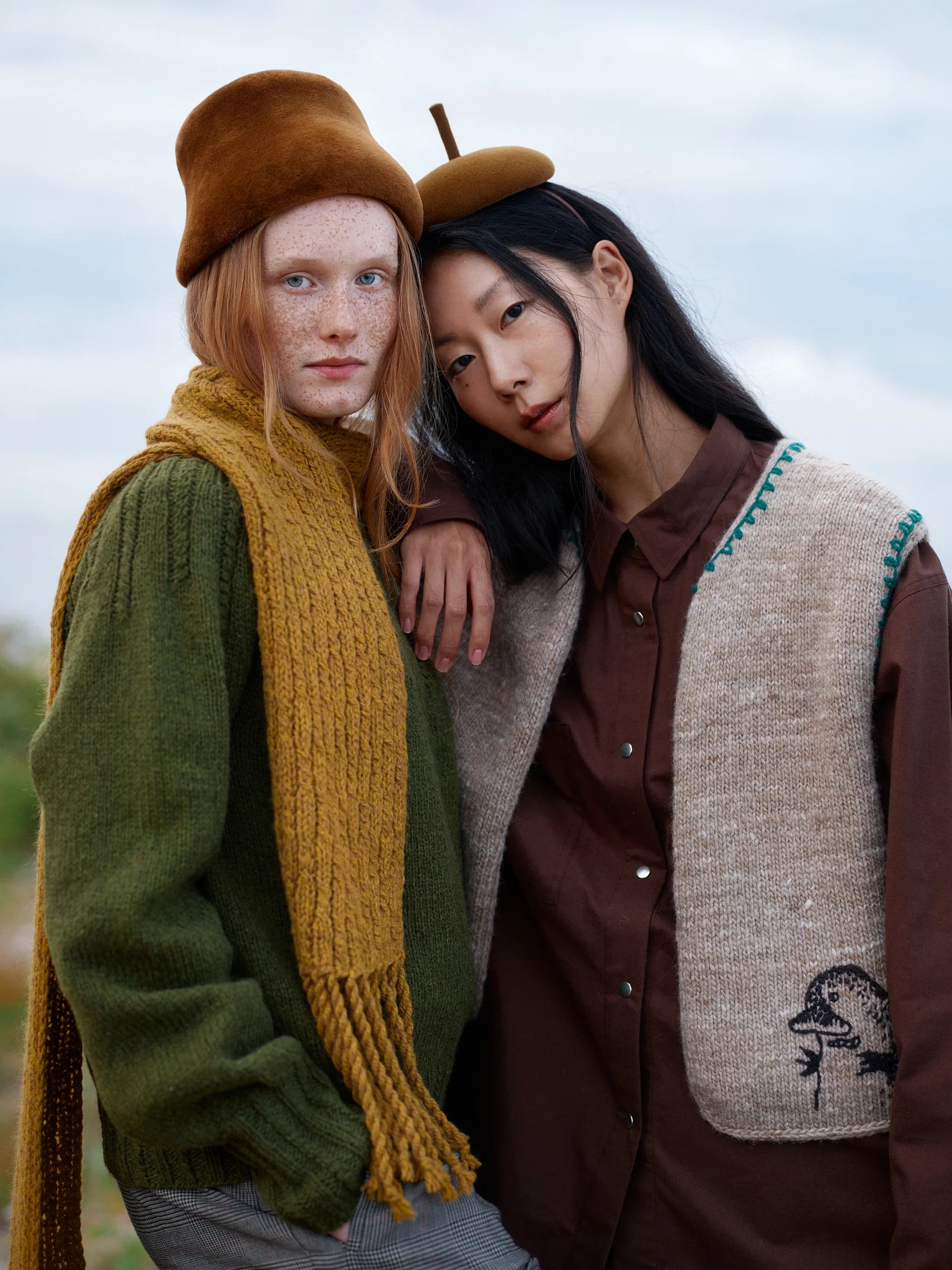Two people, one wearing a yellow knit scarf and a green knit sweater, and the other wearing a beige knit vest over a brown button-down shirt, standing outside.