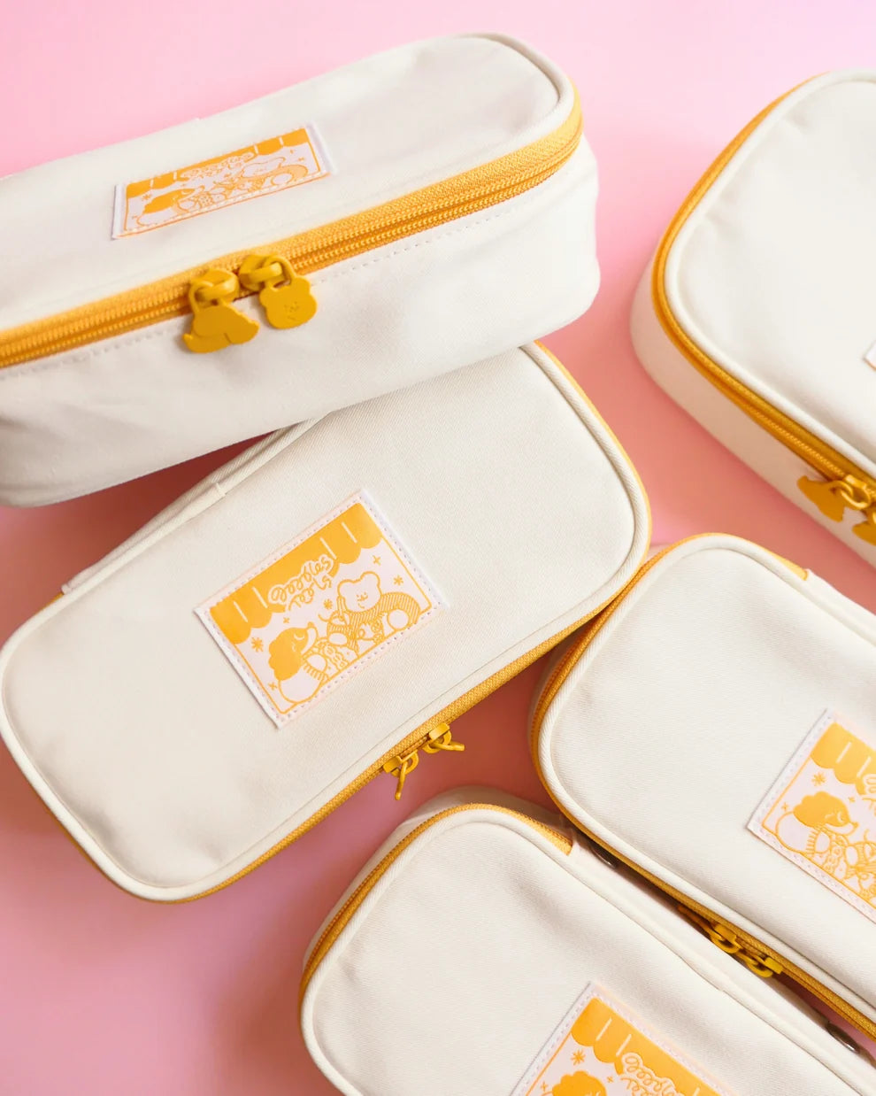 A few cream-colored fabric needle cases with mustard yellow zippers and a cute cartoon patch on the front, arranged on a solid pink background.