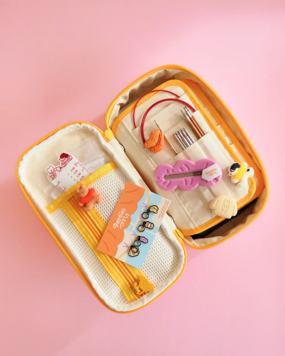 Open beige and yellow needle case with knitting needles and various small knitting accessories on a pink background
