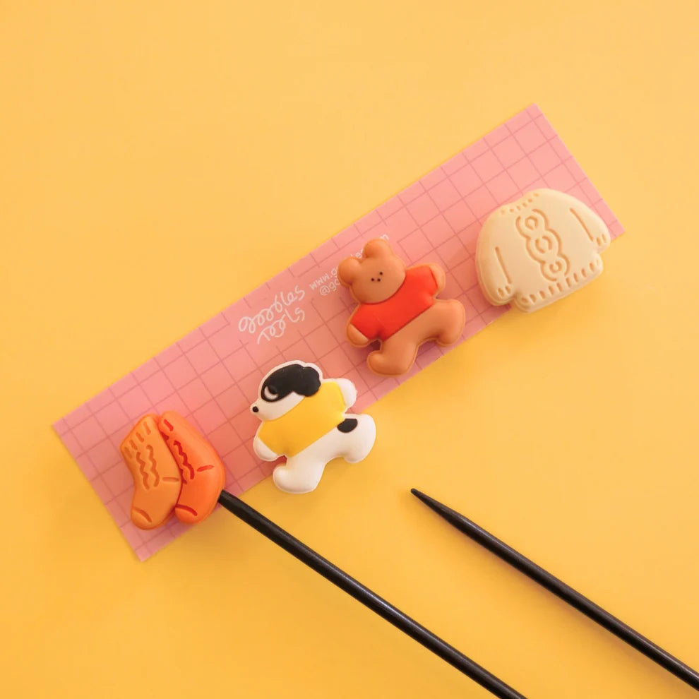 Four needle stoppers on a pink piece of cardboard against a yellow background with two knitting needles.