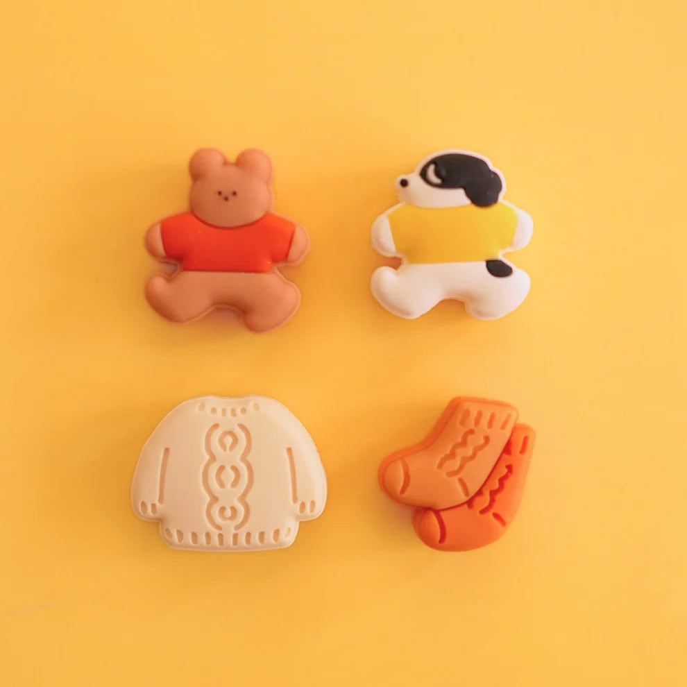 Four needle stoppers arranged on a solid yellow background: a teddy bear wearing an orange shirt, a dog wearing a yellow sweater, a cream-colored knit sweater, and a pair of orange socks.