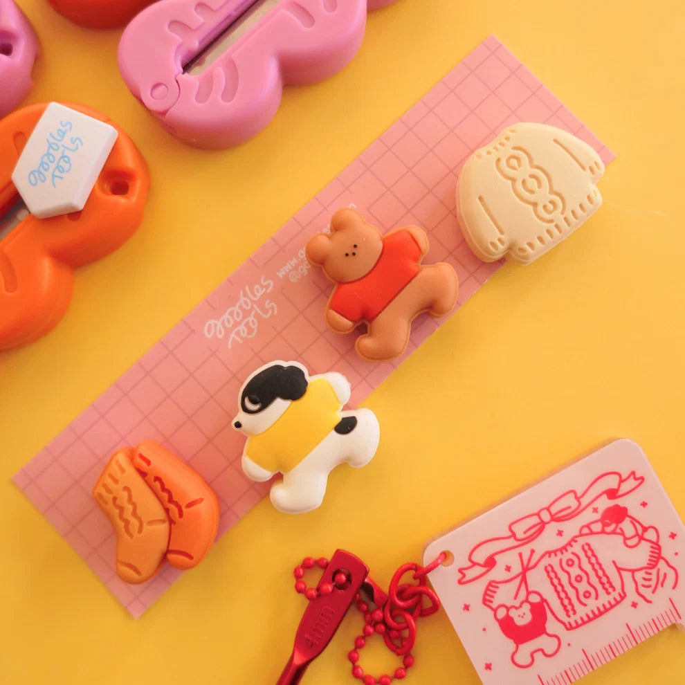 Four needle stoppers on a pink piece of cardboard against a yellow background surrounded by assorted knitting tools.