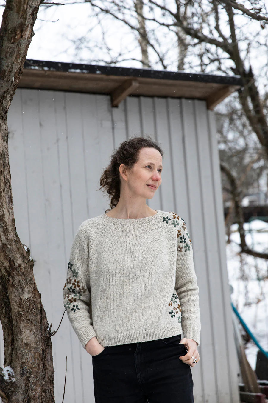 A woman wearing a light beige knit sweater with black floral embroidery on the sleeves, standing outdoors in front of a white wooden shed.