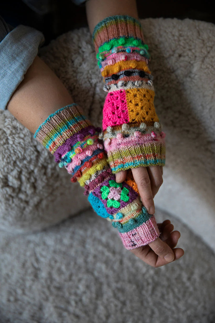 A close-up of a person's hands and forearms wearing a pair of colorful, chunky crocheted and knit fingerless wrist warmers or arm warmers, resting on a soft beige textured blanket.